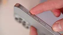 A finger presses the small Action Button on the side of an iPhone above its volume buttons.