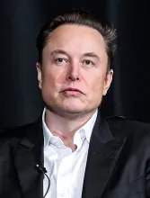 A portrait photograph of Elon Musk in a dark suit jacket and white shirt against a dark background.