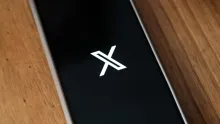 A smartphone lying on a wooden surface displays the white X logo on a black screen.