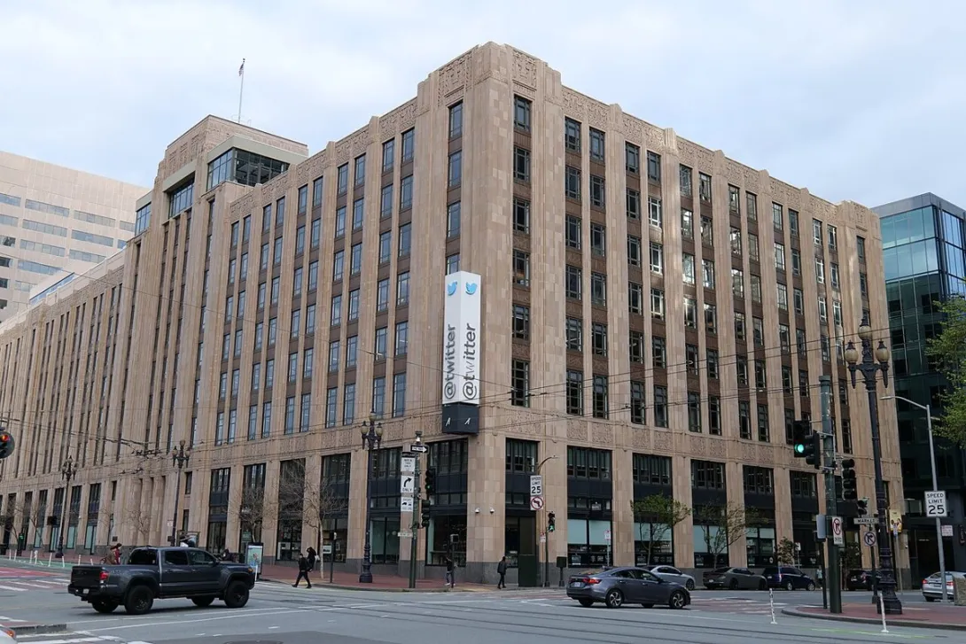 The Twitter offices in San Francisco, a large stone building with a vertical Twitter banner at a city street corner.