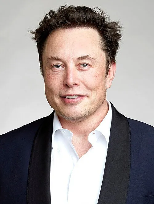 A portrait photograph of Elon Musk in a dark jacket and white shirt against a light background.