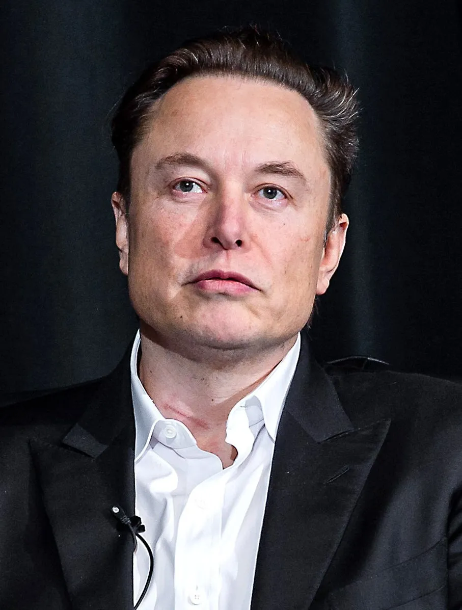 A portrait photograph of Elon Musk in a dark suit jacket and white shirt against a dark background.