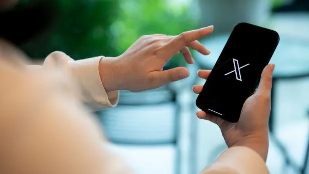 A person holds a smartphone showing the X logo while reaching to tap the screen.